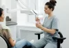 Jak bezboleśnie uzupełnić brakujące zęby w dorosłym życiu Dentystka wyjaśnia dorosłej pacjentce zabieg, trzymając model szczęki.